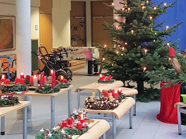 Ein Foto zeigt den geschmückten Weihnachtsbaum und die mitgebrachten Adventsgestecke und -kränze in der Pausenhalle der Brückenschule.